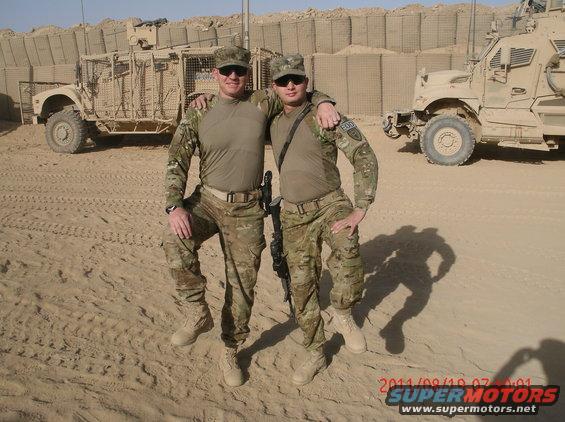 Two soldiers in camouflage uniforms and caps pose in front of military vehicles in a desert environment. One soldier has his arm around the other's shoulder