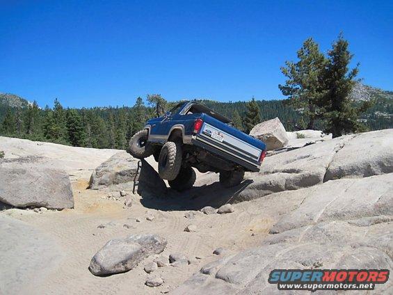 1986 Ford Bronco climbing over large rocks on a dirt trail with trees and mountains in the background