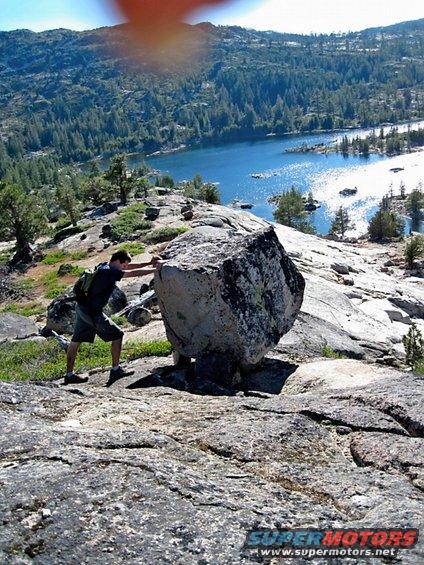 Man pushing a large boulder on a rocky hillside with a lake and trees in the background