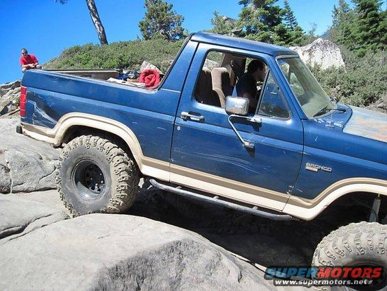 Blue Ford Bronco with tan trim, off-roading over large rocks. Visible 'BRONCO' badge on fender
