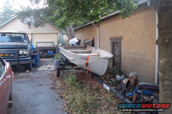 Aluminum boat on a trailer parked next to a brick building. A dark pickup truck is also visible
