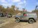 1986 Ford Bronco - A group of off-road vehicles are gathered on a dirt track with trees in the background.