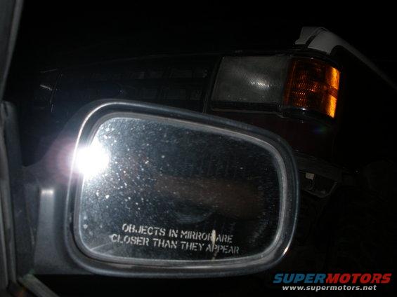 Side mirror with 'objects in mirror are closer than they appear' text, reflecting a car's headlight and orange turn signal