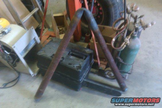 Rusty metal pipes form an A-frame over a black toolbox, with a green oxygen tank and welding torch nearby