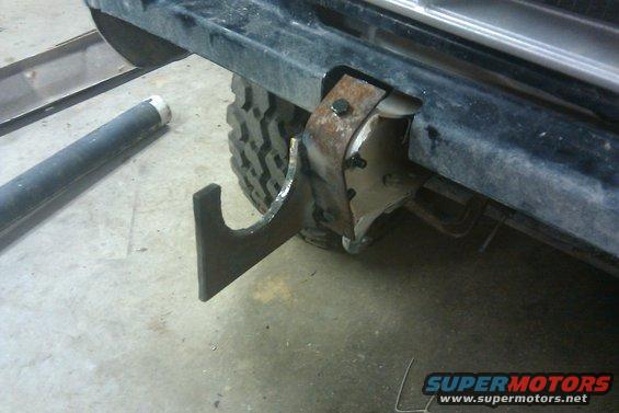 Custom metal tow hook welded to a rusty bracket on a black bumper, with a tire visible behind it