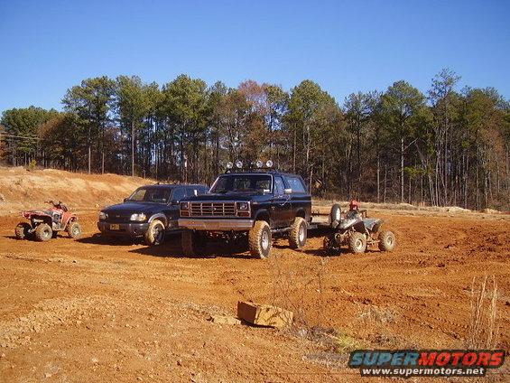 Black Ford Bronco with roof lights and a trailer carrying an ATV, parked next to a dark SUV on a dirt lot