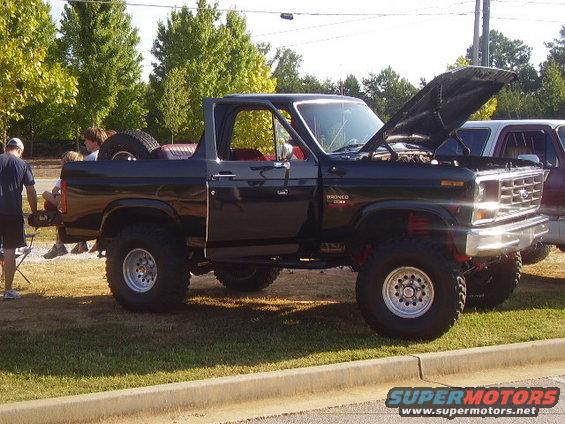 Black Ford Bronco with hood open, spare tire in back. Another vehicle is parked next to it