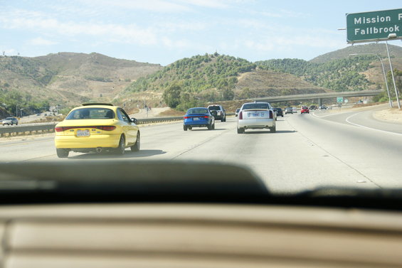 Yellow sedan driving on a sunny highway, with other cars and hills in the background. License plate is visible