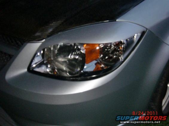 Car's front headlight and bumper area. Date and website visible