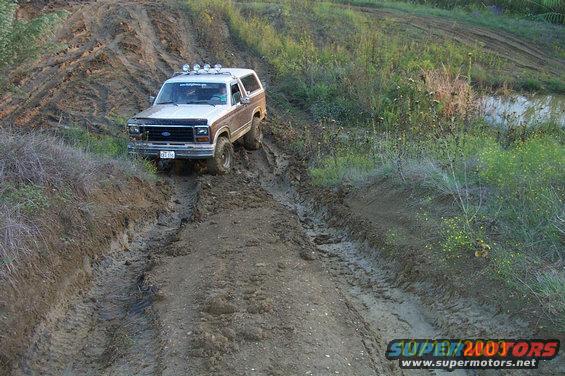 Brown and white Ford Bronco driving through mud, with roof lights