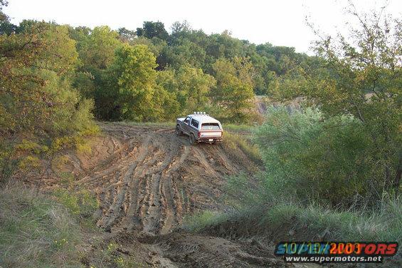 Silver 1980s Ford Bronco driving on a muddy hill with trees in background
