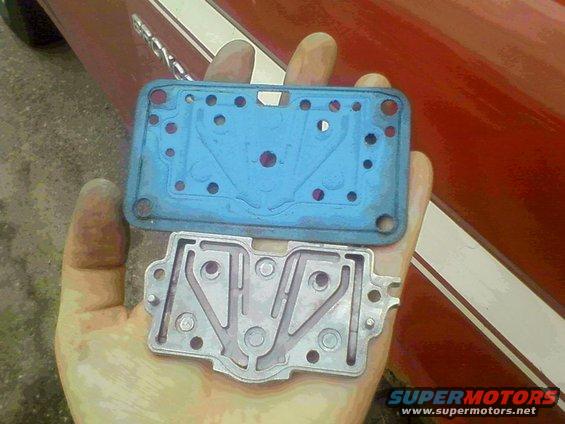 Hand holding carburetor gasket and base plate. Red vehicle in background