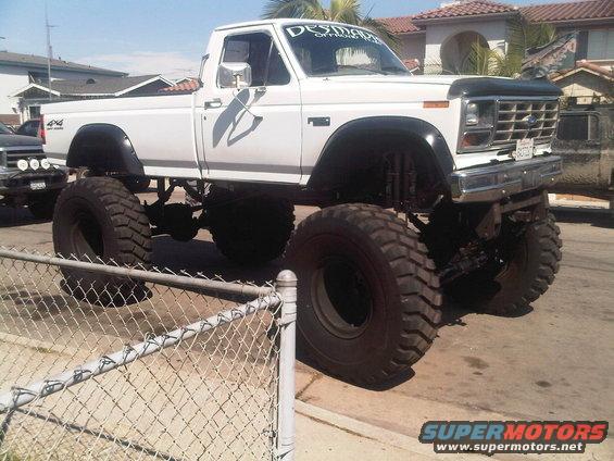 White lifted pickup truck with large tires, 4x4 badge on fender, and 'DESIMON' sticker on windshield