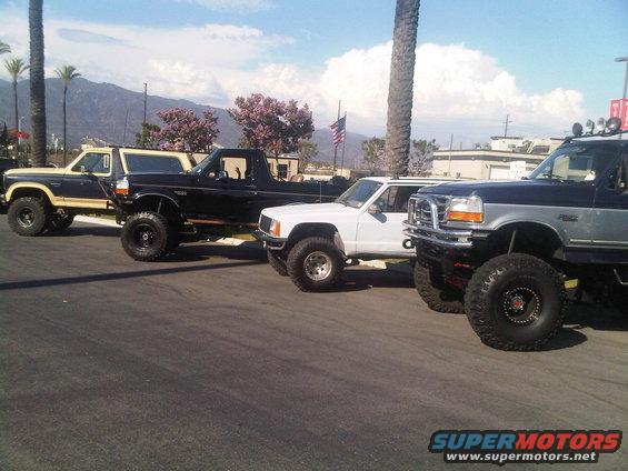 Four lifted trucks parked side-by-side, including a dark blue and tan Bronco, a black Bronco, a white Jeep Comanche, and a dark blue and silver Ford F-Series truck