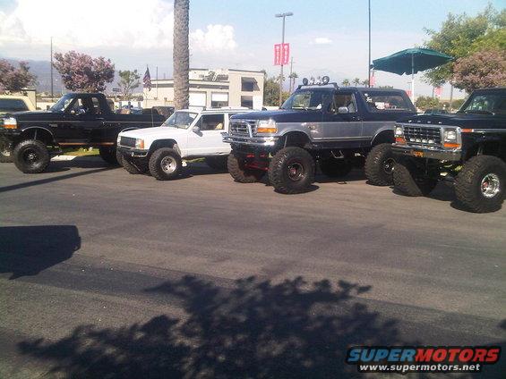 Four lifted trucks and SUVs parked in a lot. One is a white Jeep Comanche, another is a dark blue and silver Ford Bronco, and two are black Ford pickup trucks