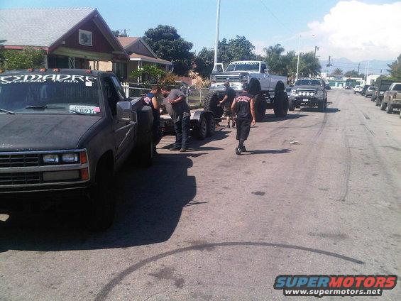 Several modified pickup trucks and trailers parked on a street, with people gathered around one