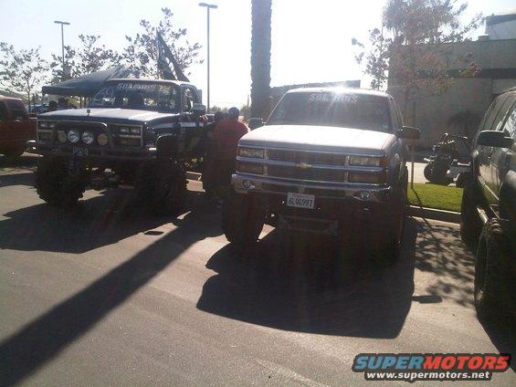 Two lifted pickup trucks parked outside, one with a flag on top