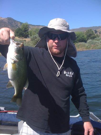guy in hat and sunglasses holding a largemouth bass, standing in a boat on a lake