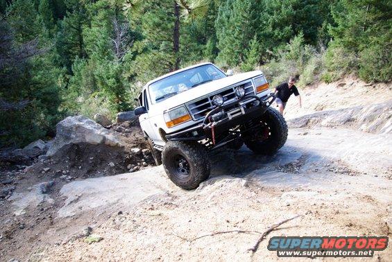 White SUV with off-road modifications driving on a rocky trail. Person walking behind it