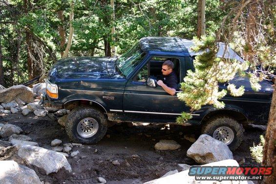 Dark green Ford Bronco navigating a rocky, wooded trail. Driver wears sunglasses