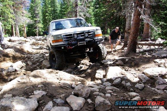 White 1995 Ford Bronco navigating a steep, rocky trail with a person walking behind it