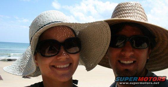 Two people wearing wide-brimmed hats and sunglasses smile at the camera on a sunny beach with the ocean in the background