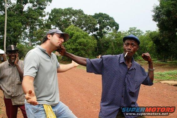 Three men stand on a dirt road with trees in the background; one man is being slapped