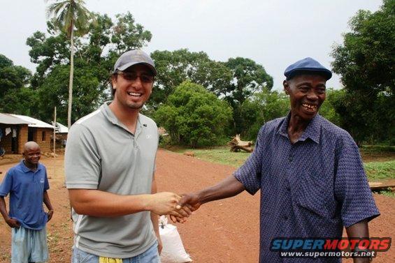 Two men shaking hands in a rural village with a palm tree and simple dwellings