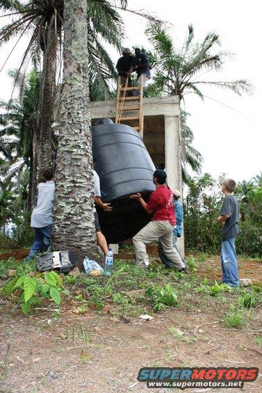Several people are struggling to move a large black water tank next to a palm tree and a concrete structure. Some are on a ladder, others are pushing