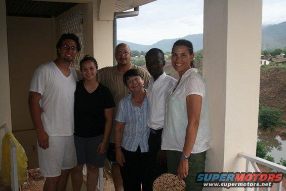 Six people stand on a covered porch overlooking a valley with a body of water