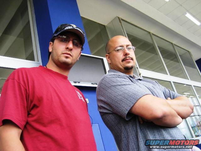 Two men stand in front of a building with large windows and blue accents. One wears a red shirt and a baseball cap, the other a grey polo