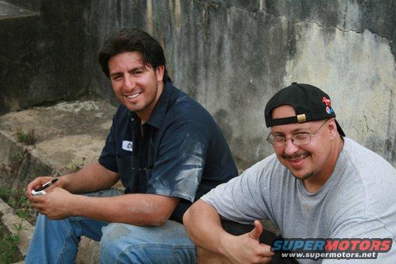 Two men sitting on concrete steps outdoors. One wears a blue work shirt, the other a grey t-shirt and baseball cap