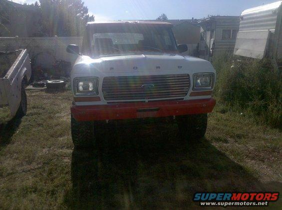 1979 Ford Bronco front view, red bumper, chrome grille. Another vehicle part is visible to the left
