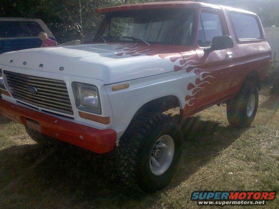 Ford Bronco with white and red paint, flame decals, and large tires. Another vehicle is partially visible to the left