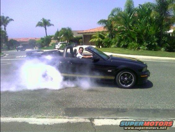 Black convertible sports car doing a burnout, side view. Appears to be a 2005 Ford Mustang