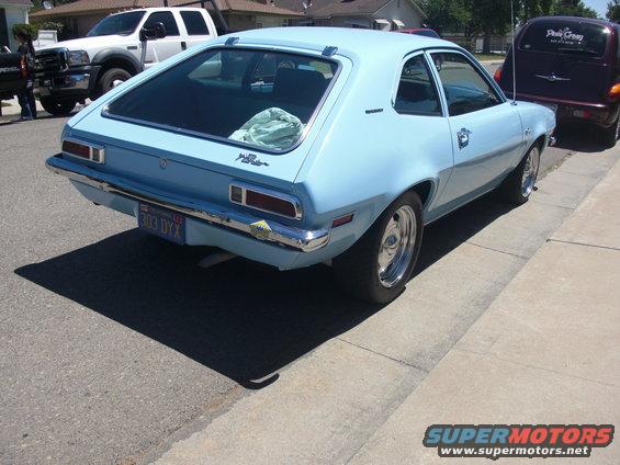 light blue 1972 Ford Pinto hatchback parked on street, rear view. White pickup truck and dark minivan also visible