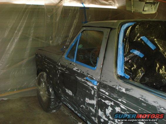 Pickup truck body covered in primer and blue masking tape, inside a plastic-covered booth