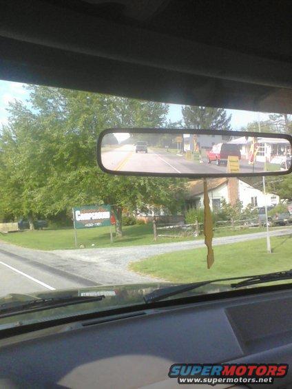 View from inside a vehicle, looking out the windshield and through the rearview mirror at a road with trees and houses. A red van and another vehicle are visible in the mirror