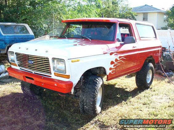 White and red Ford Bronco with flame decals, lifted with big tires. Another blue Bronco is parked behind it