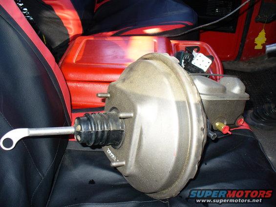 Brake booster and master cylinder assembly on black and red seats, with a red plastic container behind it