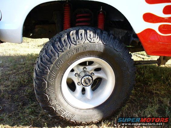 Close-up of a large off-road tire on a silver rim, showing red shocks and springs. Red and white flame decals are visible on the vehicle body