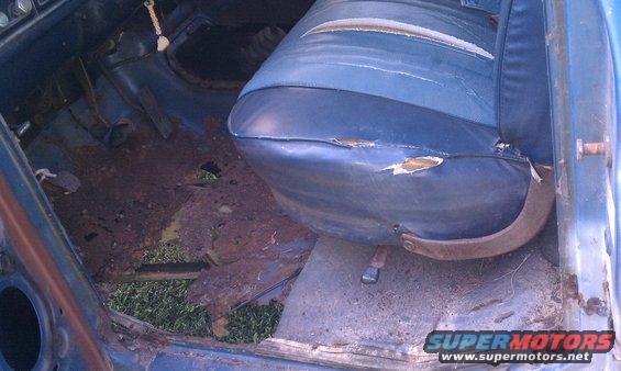 Interior view of a vehicle with heavily rusted floorboards and torn blue upholstery on the driver's seat