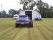 1992 Ford Bronco - A blue pickup truck with massive tires is parked in a grassy field in front of a metal building.