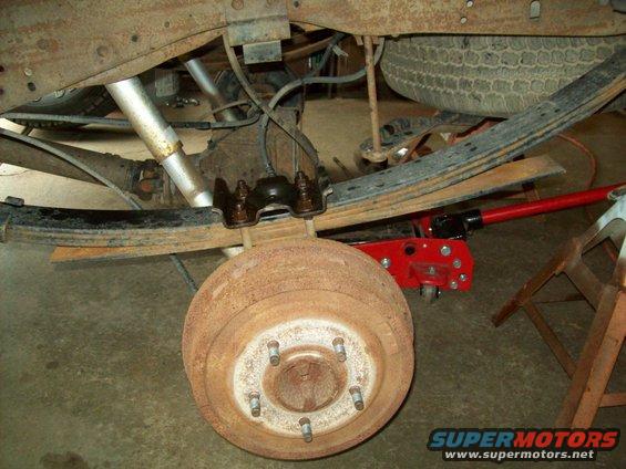 Rusty rear drum brake assembly and leaf springs on a truck, with a red hydraulic jack underneath