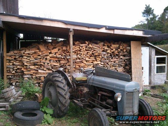 Old gray tractor parked in front of a woodpile next to a shed