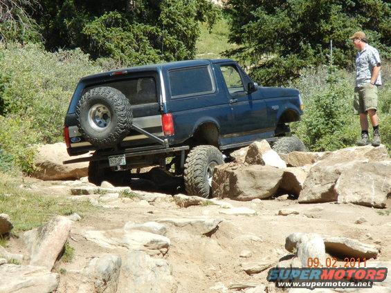 Dark blue Ford Bronco off-roading over rocks, spare tire mounted on rear