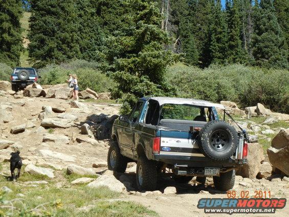 Ford Bronco on a rocky trail, another SUV behind it. Dog runs ahead