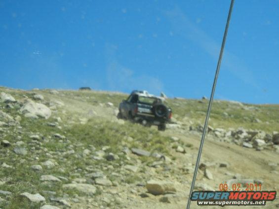 1996 Ford Bronco driving up a rocky, grassy hill under a clear blue sky