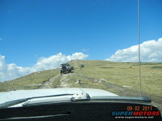 Several SUVs driving up a rocky dirt road on a grassy hill under a blue sky with clouds