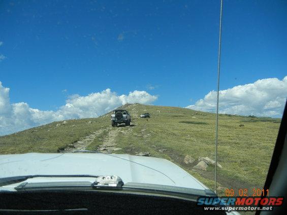 White Ford Bronco driving on a dirt road up a grassy hill under a blue sky with clouds